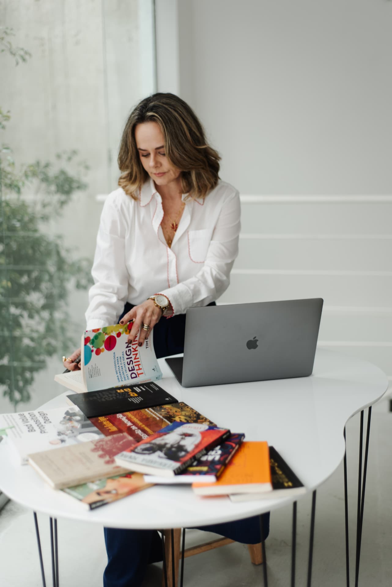 Mulher de pé diante de uma mesa branca com livros e notebook, organizando materiais de trabalho