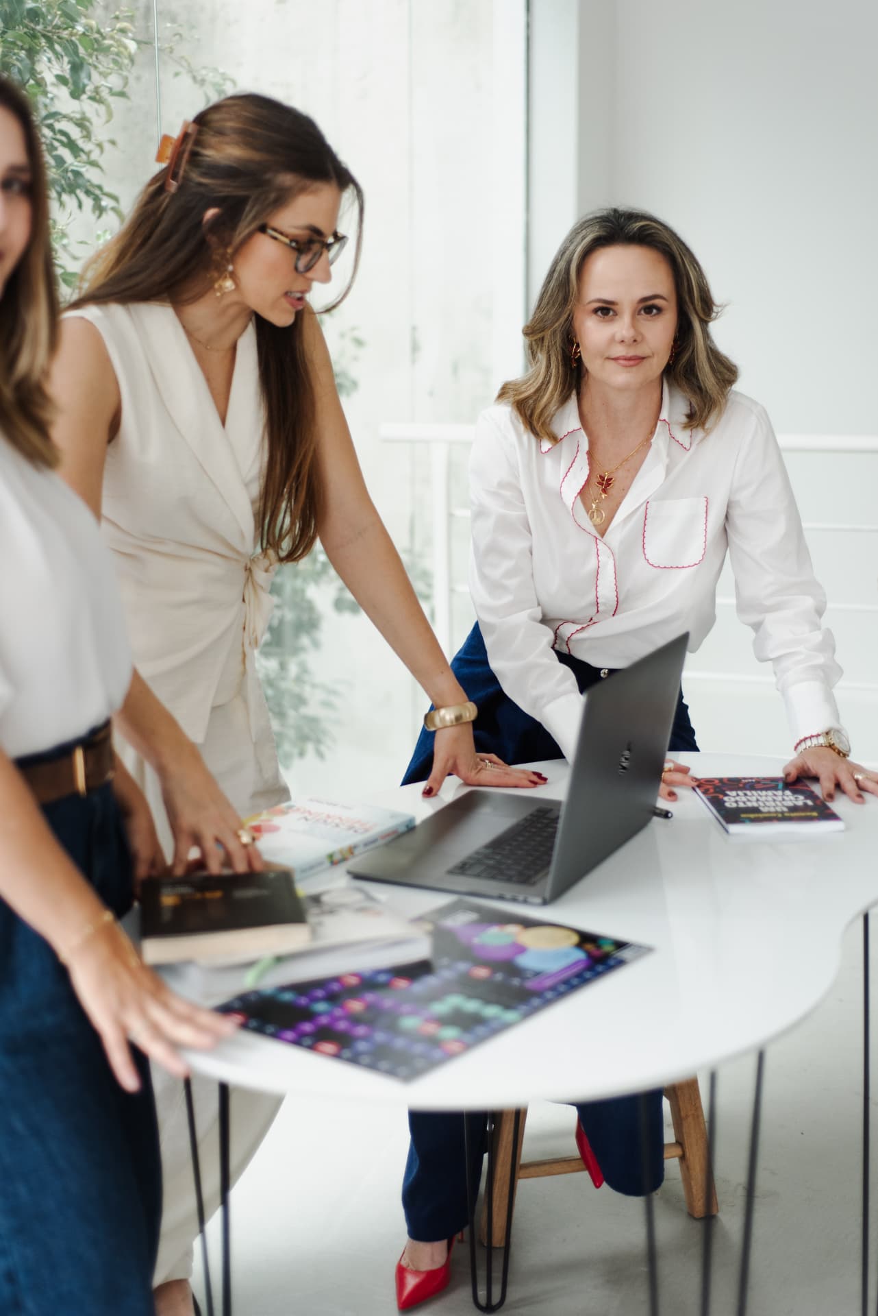 Equipe de mulheres em reunião de trabalho ao redor de uma mesa clara com livros e notebook